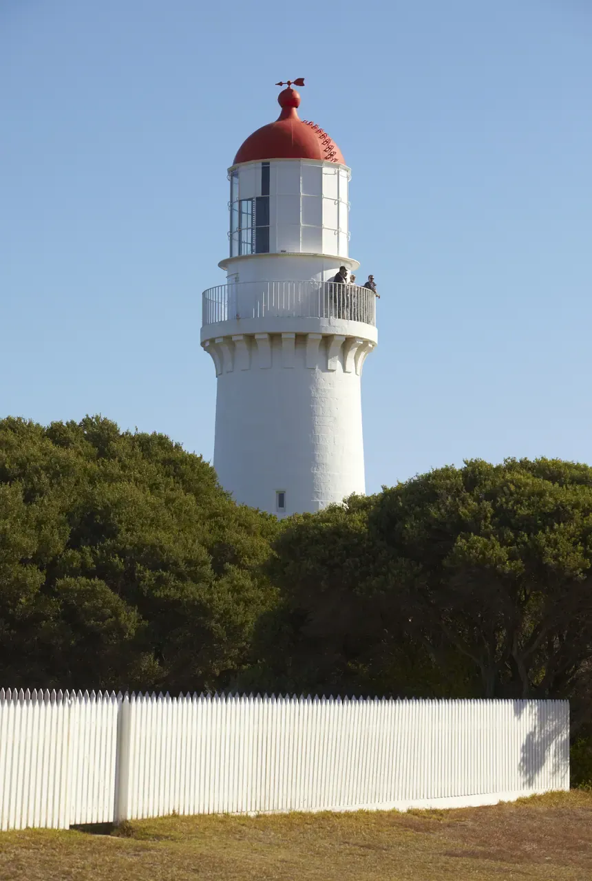 Cape Schanck Lighthouse, a white tower with a red dome, rises above trees and a white picket fence, with visitors on its balcony