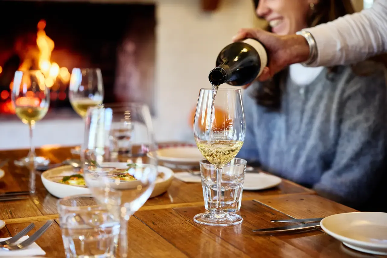 White wine is poured into a glass at a table set with a shared plate, an open fire glowing in the background