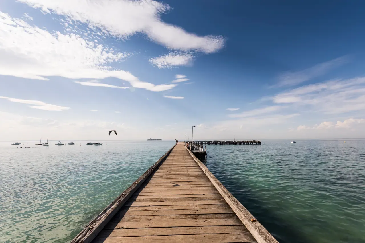 Looking along the weathered boards of Portsea Pier towards clear green water, moored boats and a passing ship