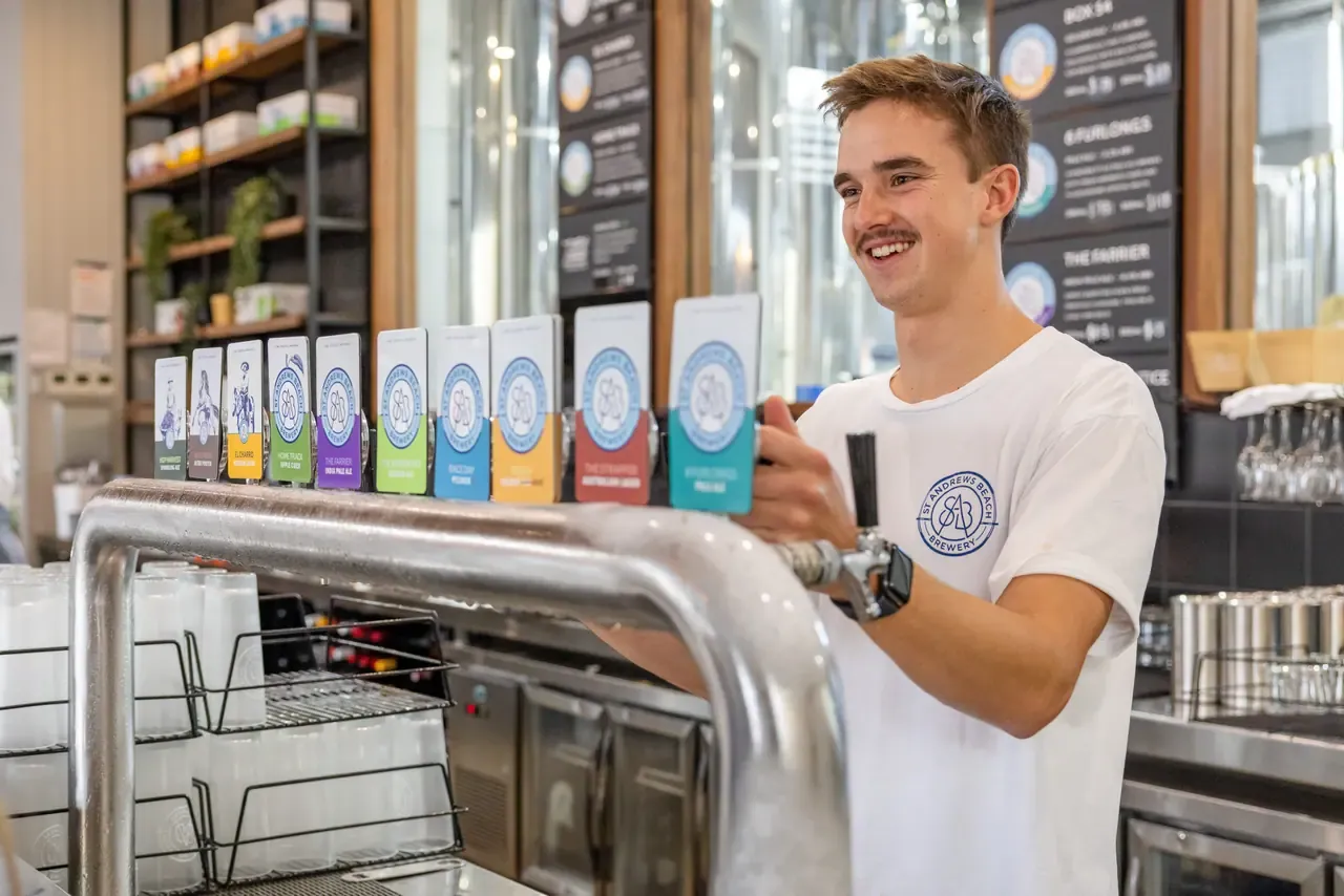 A bartender smiles as he pulls a beer from a long row of colourful tap badges at St Andrews Beach Brewery