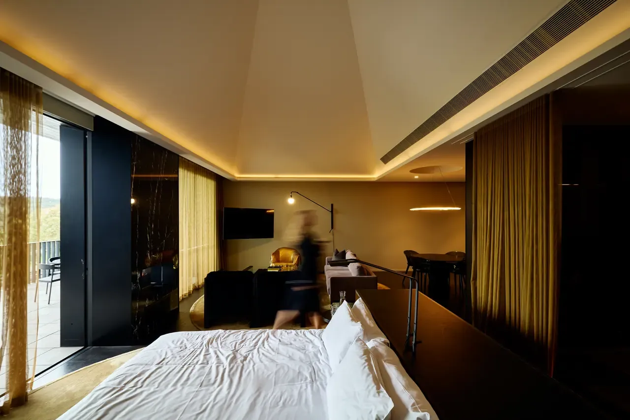 A suite at Jackalope Hotel with a white bed, gold sheer curtains, lounge area and a pitched ceiling lit by warm cove lighting