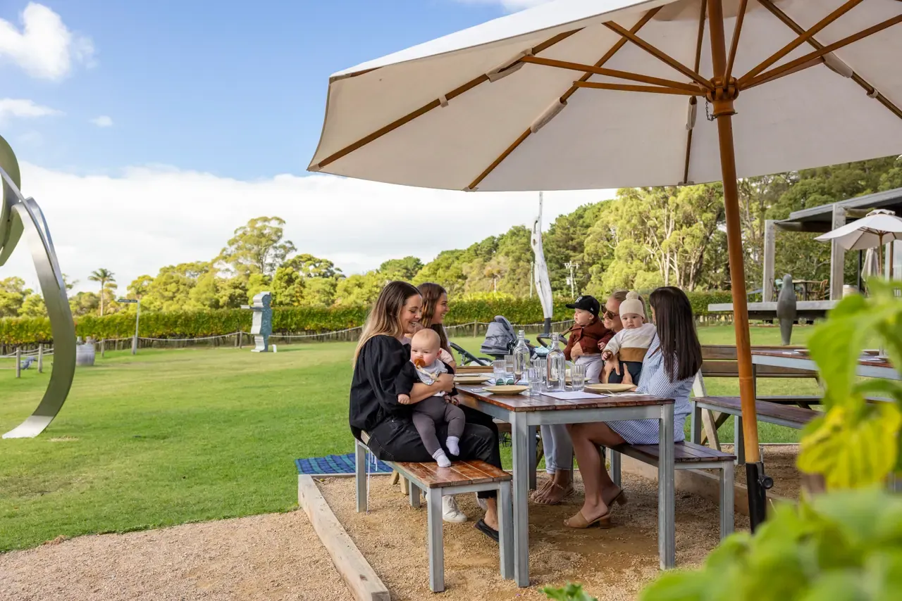 A family with babies shares lunch at a picnic table under a large umbrella on the lawn at Montalto, with sculptures nearby