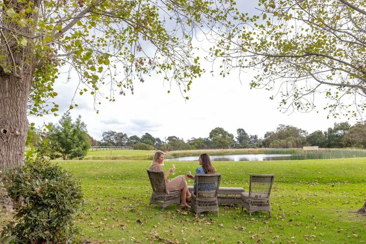 Two people sharing a glass of wine in wicker chairs on the lawn at Trofeo Estate, beneath a tree near a dam