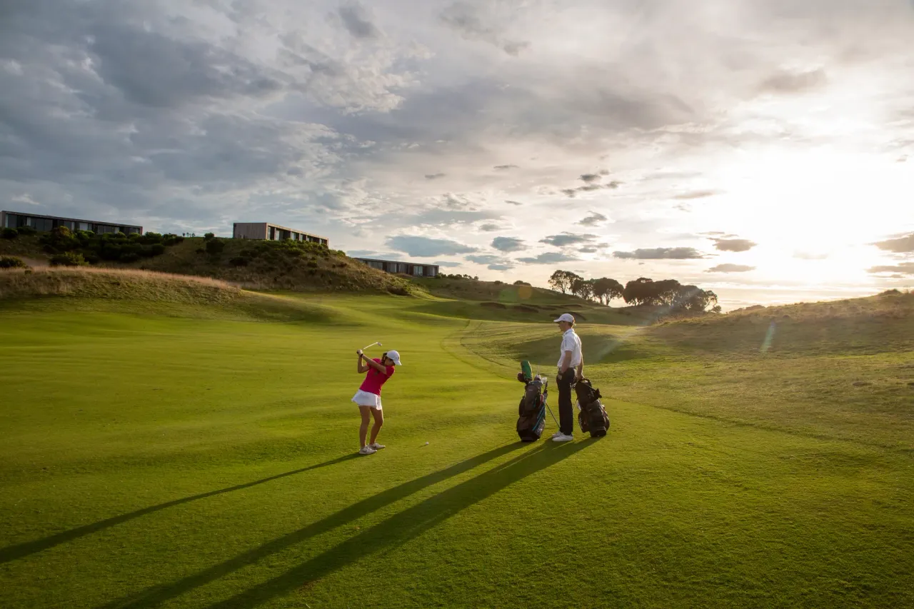 A golfer plays a fairway shot at The Dunes Golf Links as a partner stands by the bags, long shadows under a bright cloudy sky