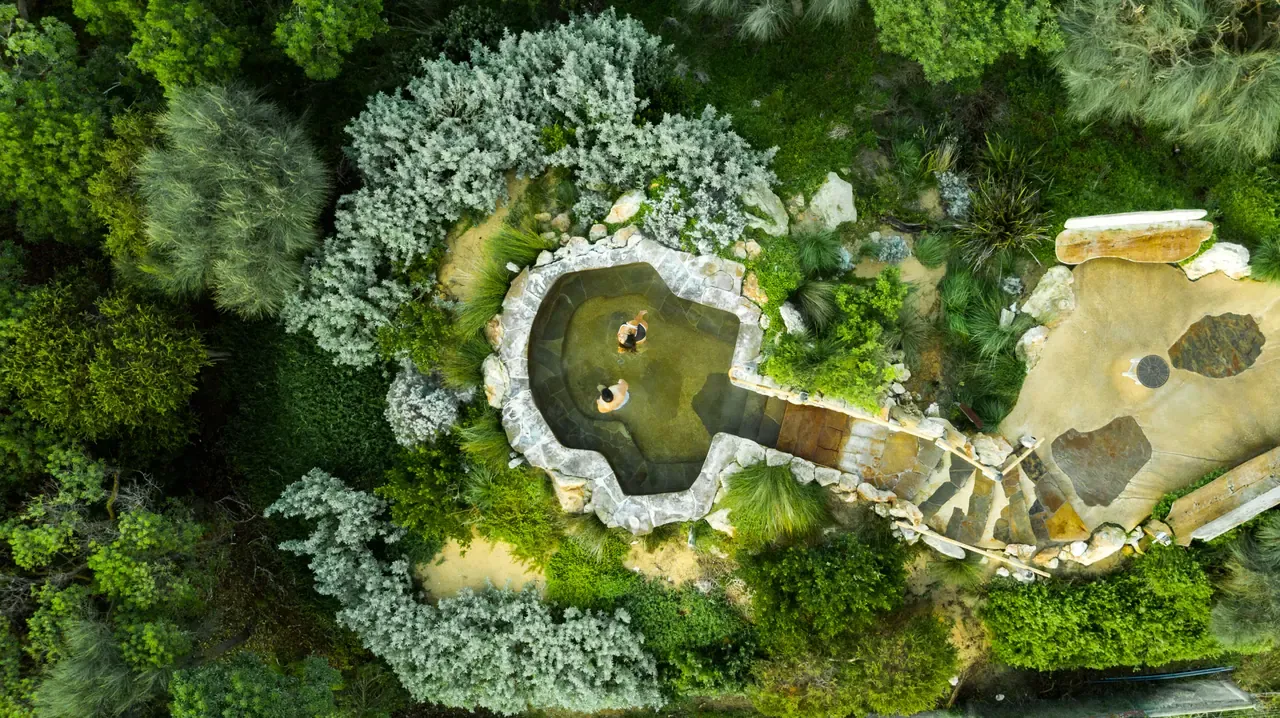 Overhead view of two bathers in a rock-edged thermal pool surrounded by silver and green native plantings at Peninsula Hot Springs
