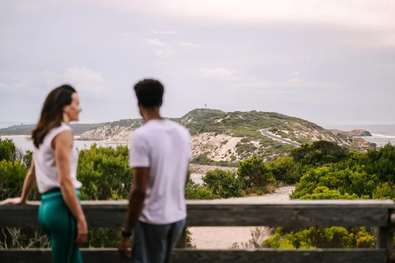Two walkers at a timber lookout rail gaze towards the scrub-covered headland and winding road leading to Fort Nepean