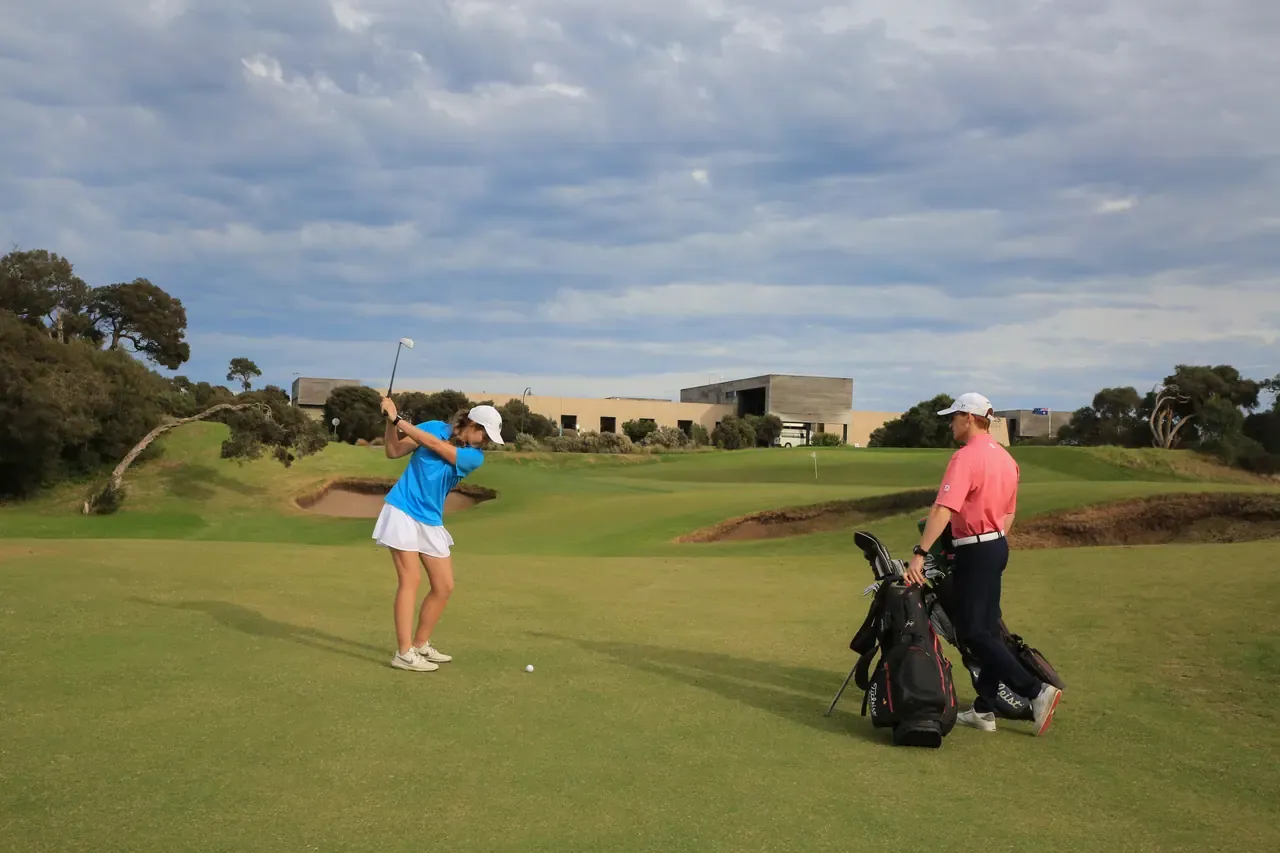A golfer takes a swing near a bunker at Moonah Links while a partner stands by the bags, the clubhouse on the ridge behind