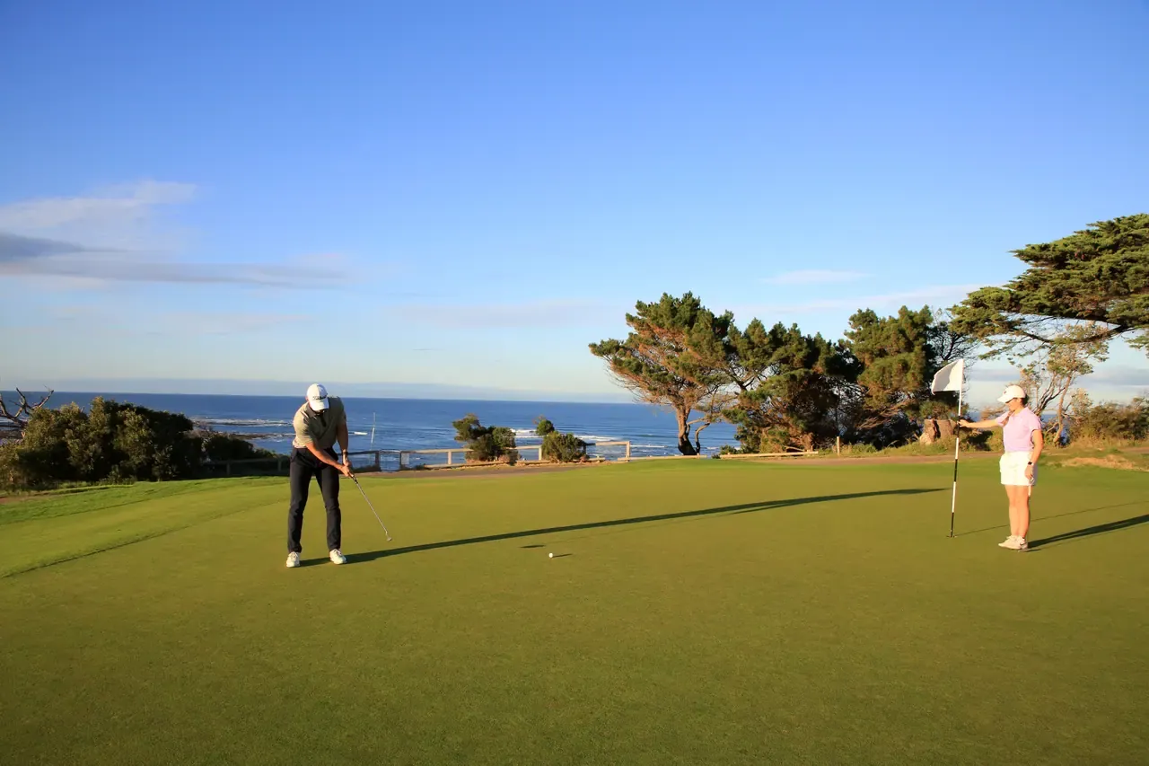 A golfer putts on a seaside green at Flinders Golf Course as a partner tends the flag, cypress trees and blue ocean behind