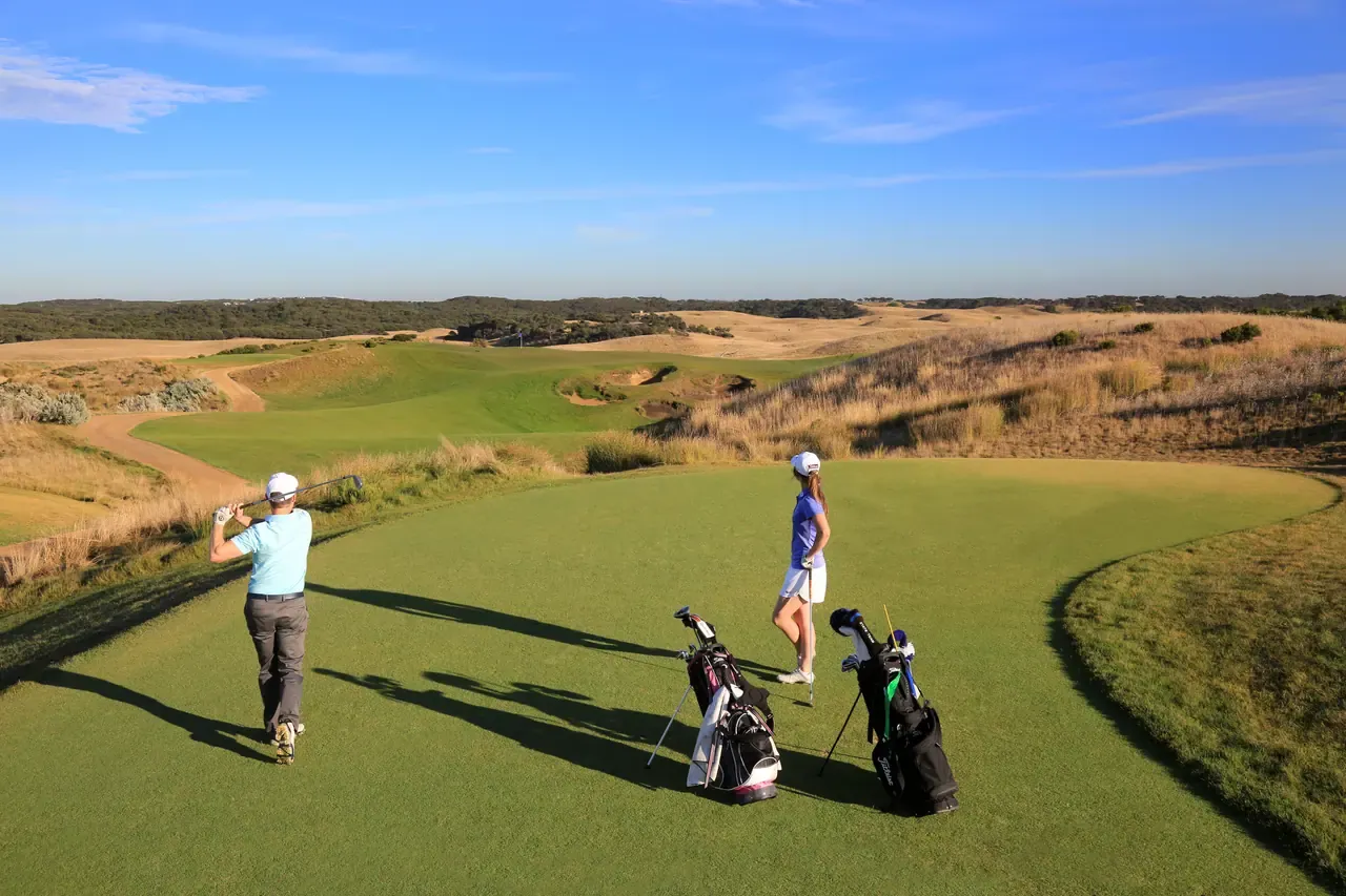 A golfer drives from a tee at The National Golf Club while a partner stands by the bags, the fairway winding through dune grass