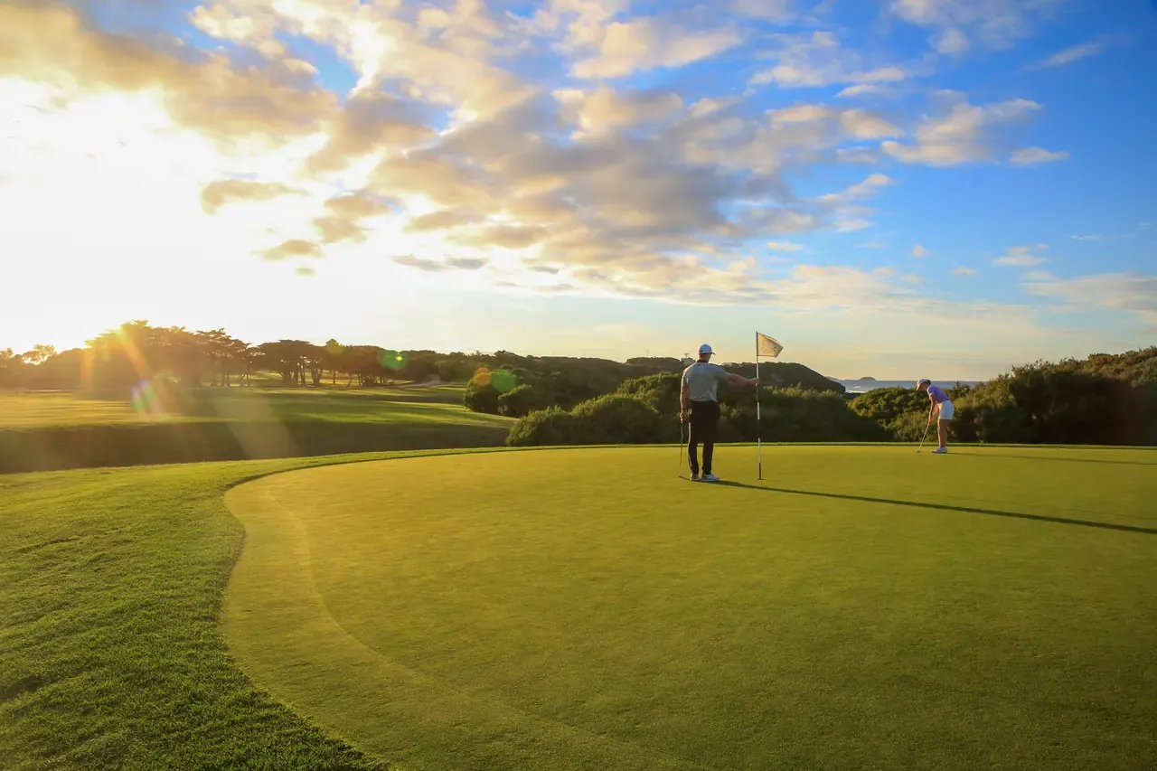 Two golfers on a green at Flinders Golf Course with the sun low behind scattered cloud and a glimpse of sea past the scrub