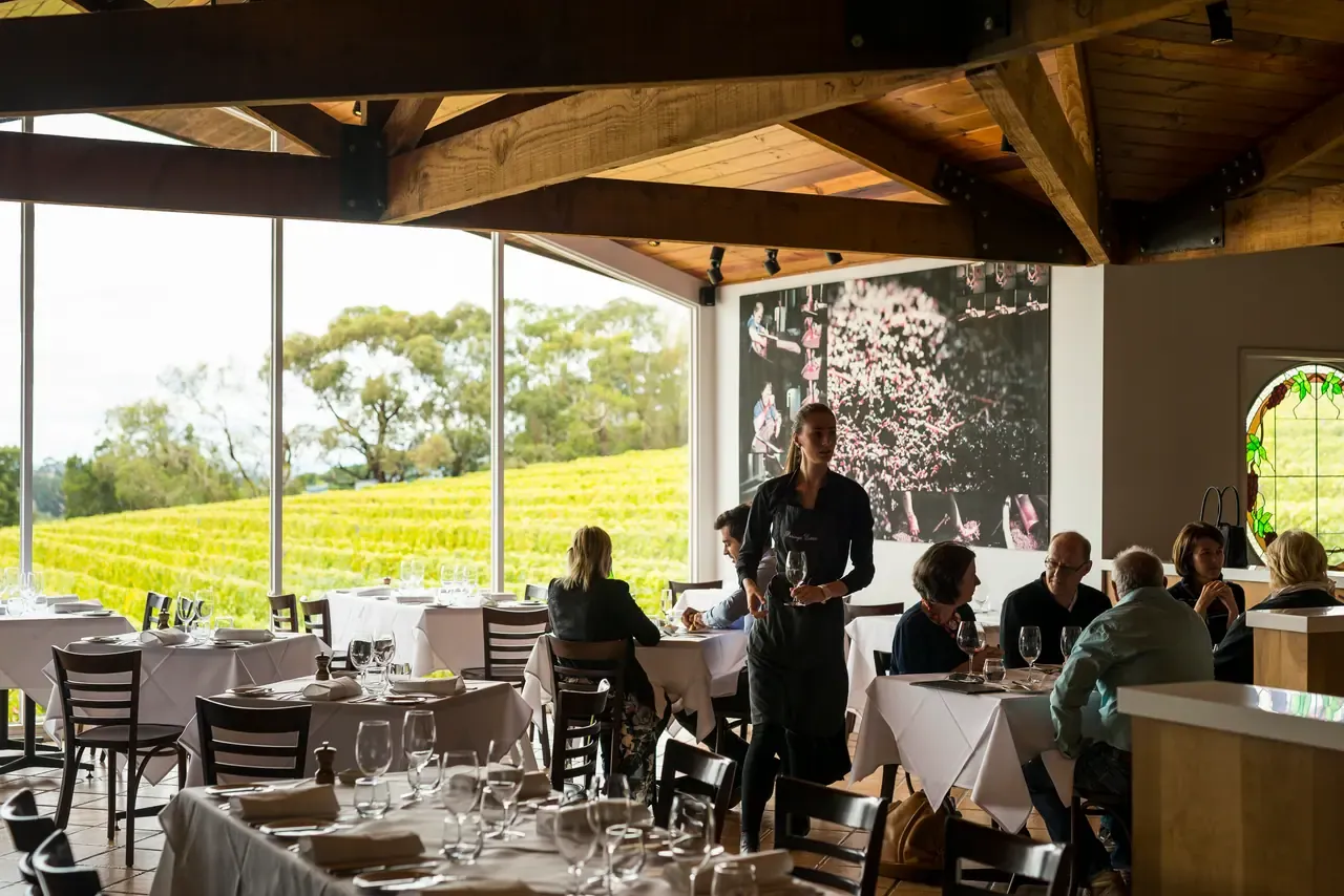 A waiter carrying wine through the Paringa Estate dining room, with tall windows over autumn-gold vineyard rows