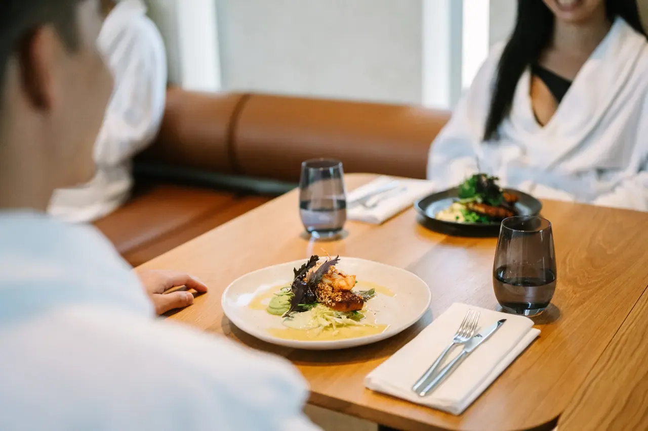 Plated fish with leafy greens on a sauce on a timber table, with water glasses and cutlery and diners in white robes behind