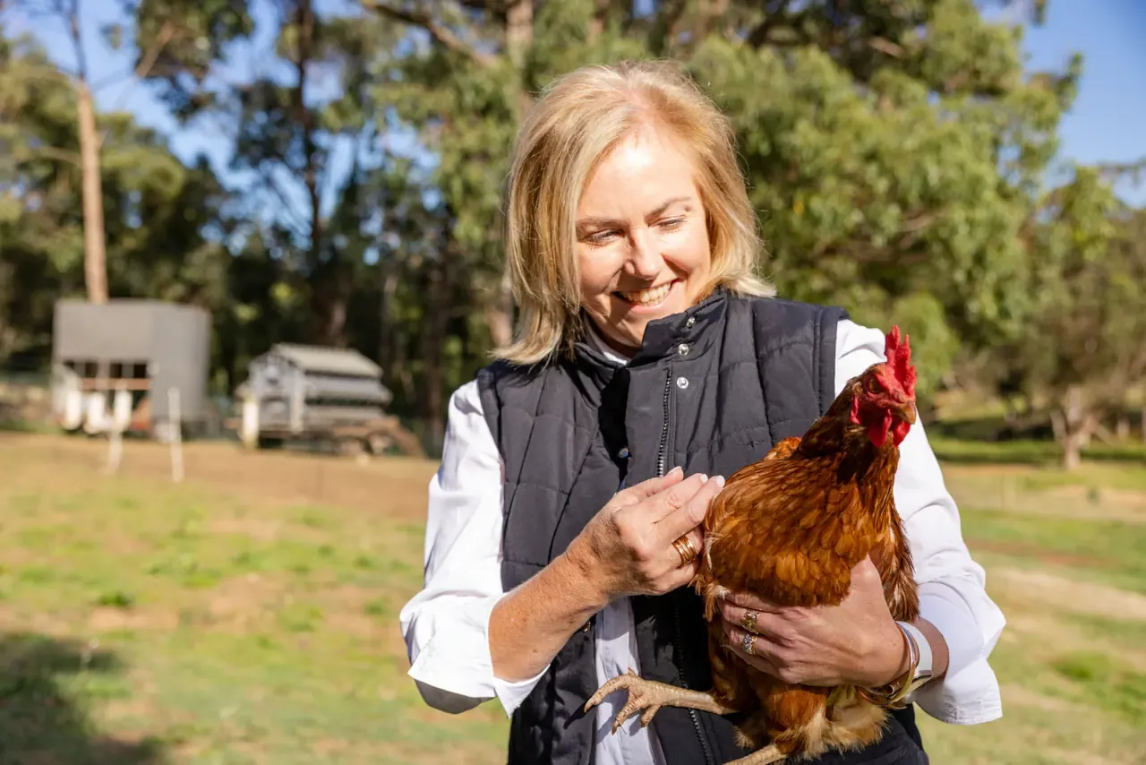 A smiling woman in a quilted vest cradles a brown hen in a sunny paddock, with chicken coops in the background