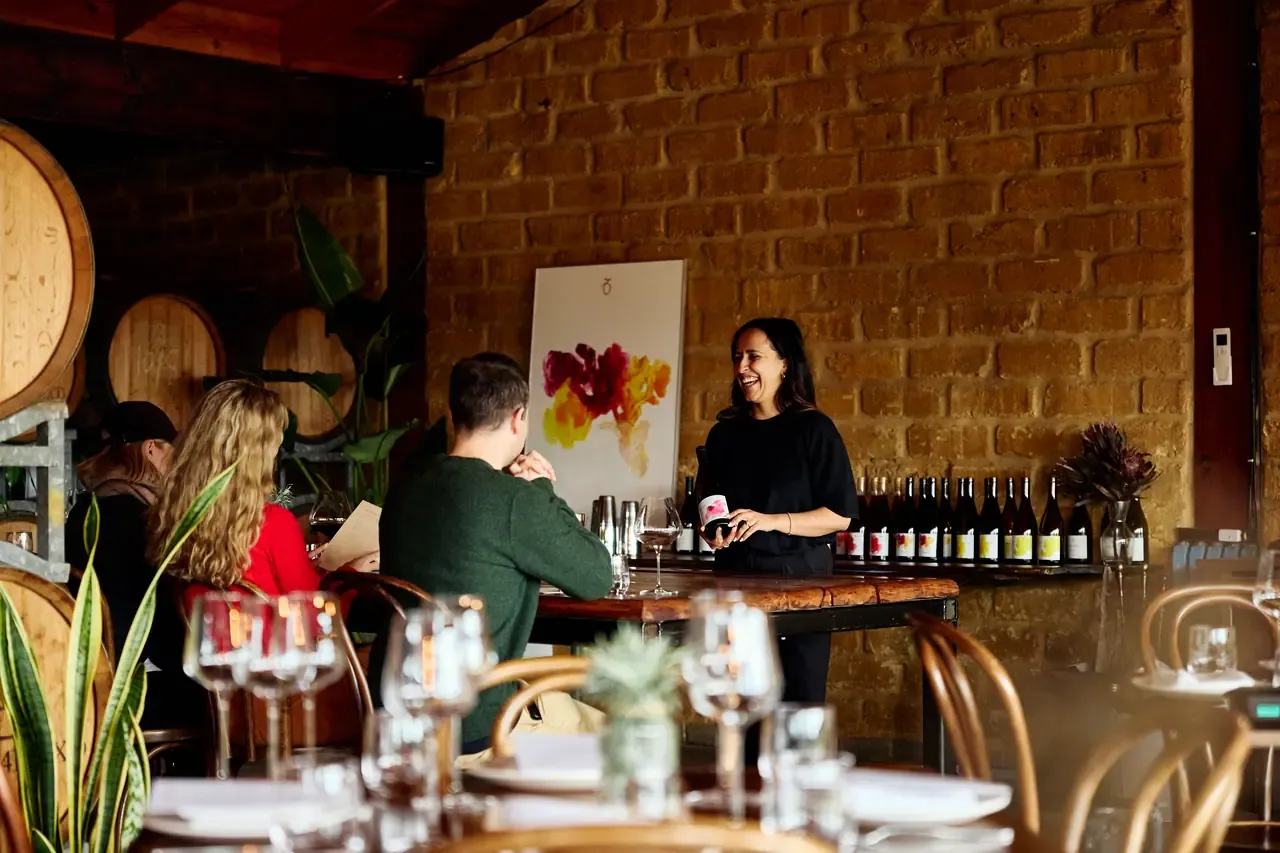 A host leading a tasting beside rows of wine bottles against a mudbrick wall at Avani Winery, barrels at left