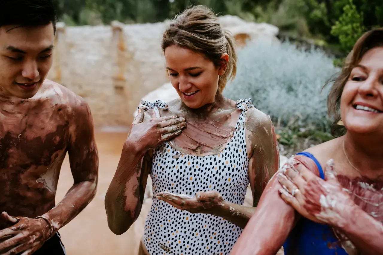 Three laughing guests with mineral mud smeared over their arms, shoulders and chests beside a sandstone wall and native plants