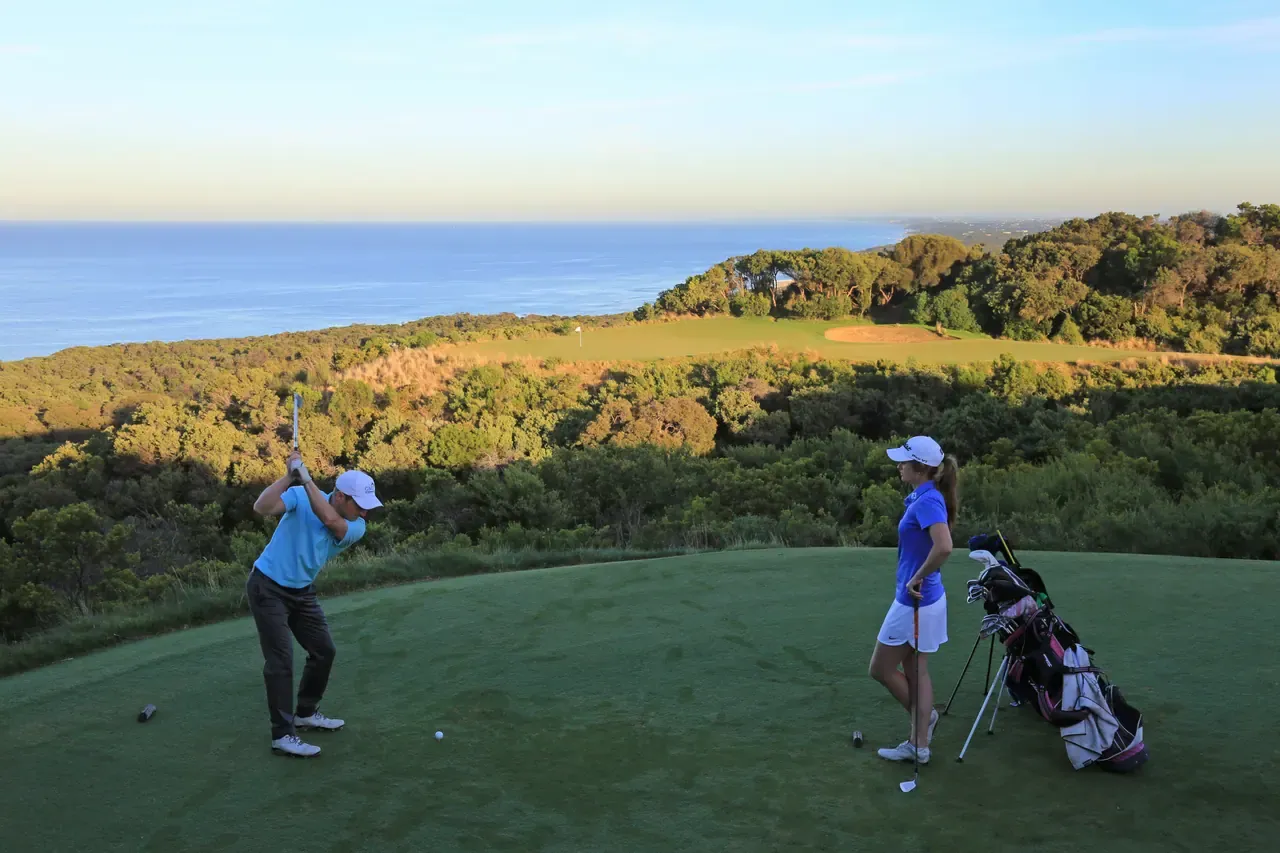 A golfer tees off high above the coast at The National Golf Club, a partner by the bags and Bass Strait stretching to the horizon