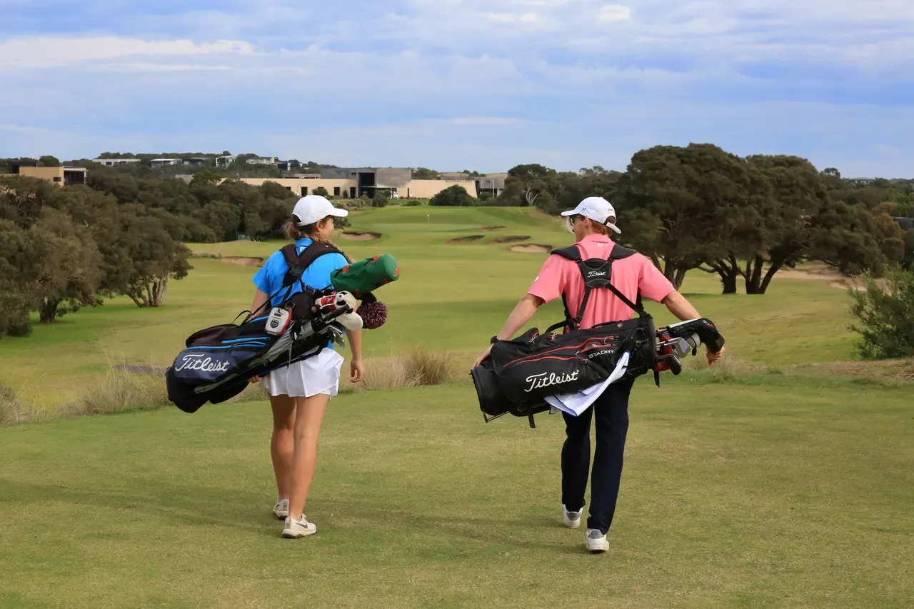 Two golfers carry Titleist bags down a fairway at Moonah Links, with bunkers and the resort buildings among trees ahead