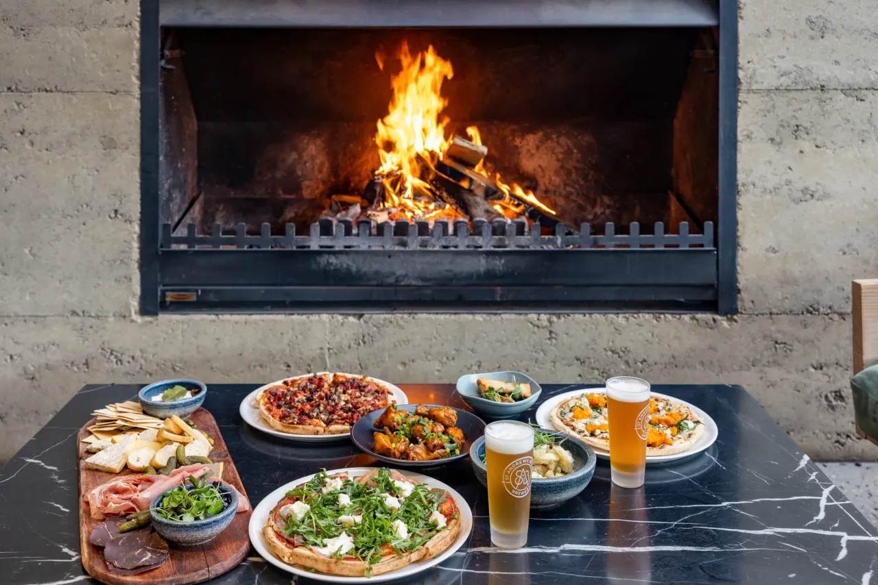 Pizzas, a grazing board, fried chicken and two pints of beer on a marble table before a blazing open fireplace