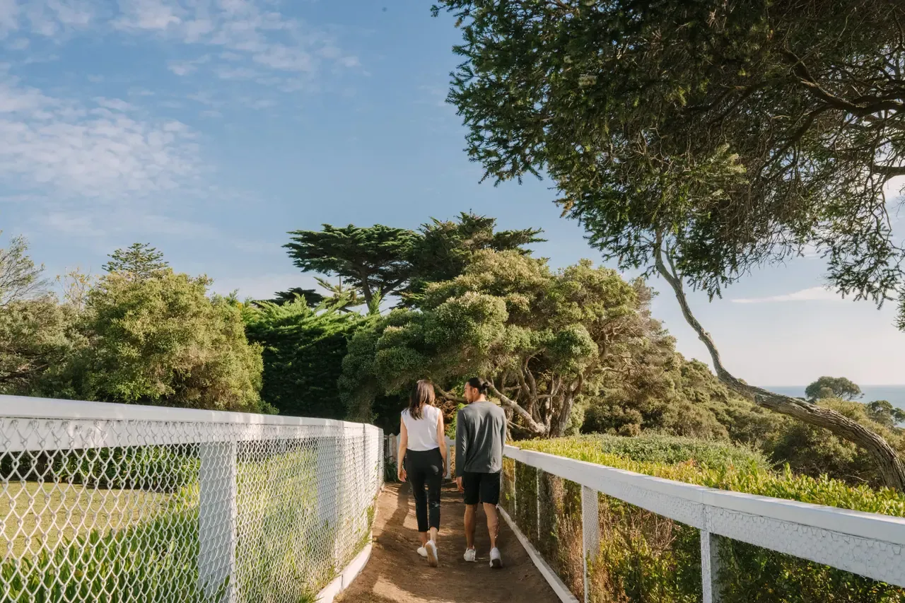 Two people walk away along a narrow path between white fences on Millionaire's Walk, shaded by twisting coastal trees