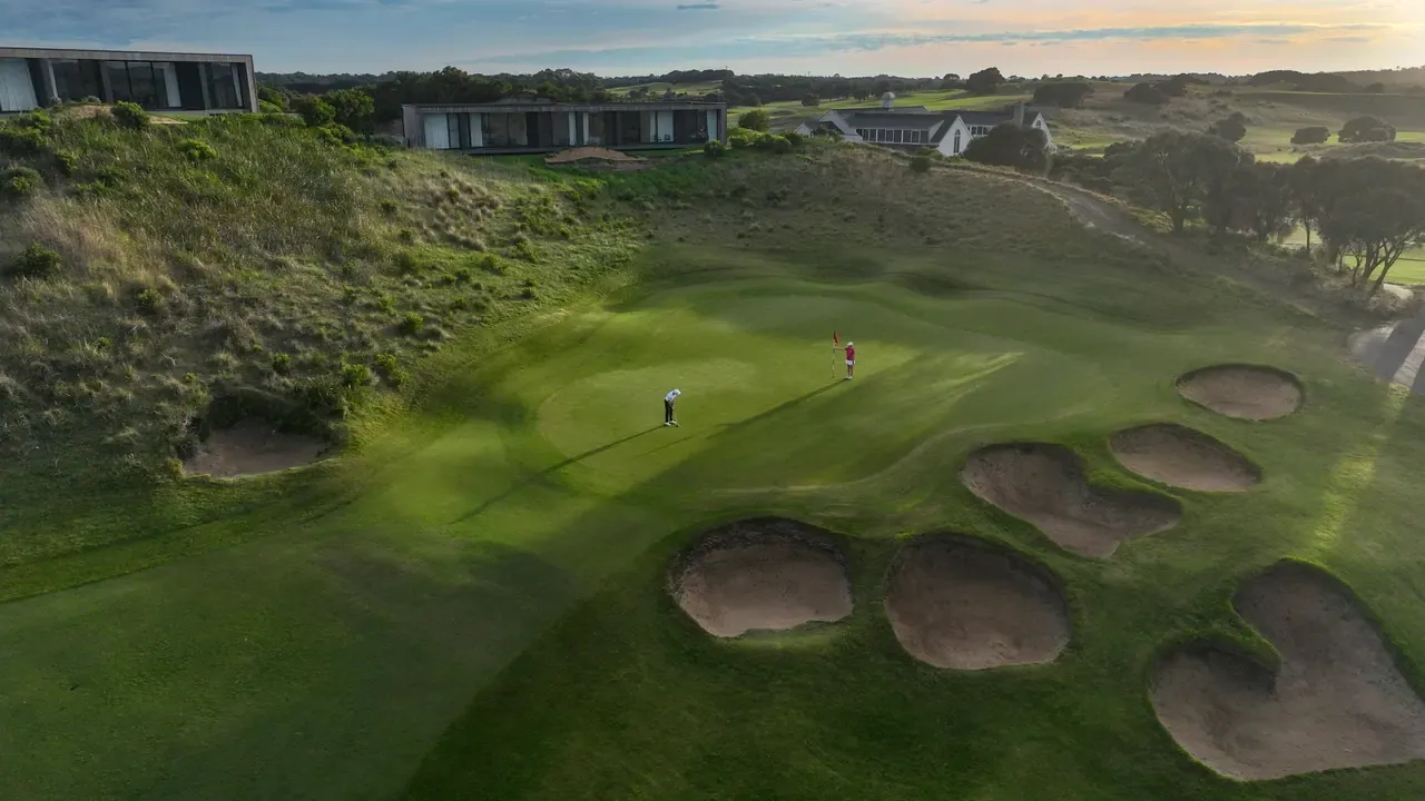 Aerial view of a green ringed by round sand bunkers at The Dunes Golf Links, two golfers putting below modern buildings on the rise