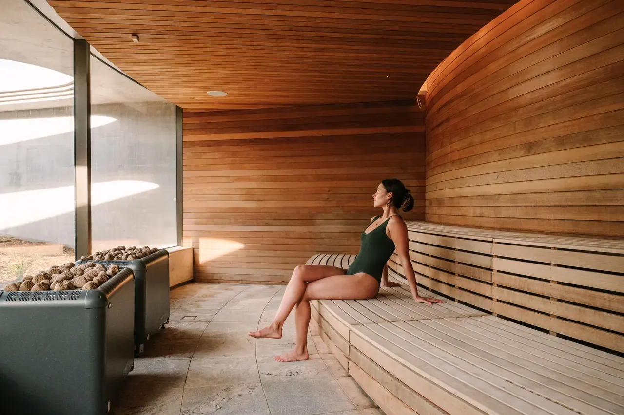 Guest in a swimsuit sits on a slatted timber bench in a curved timber-lined sauna, with heated sauna stones in the foreground