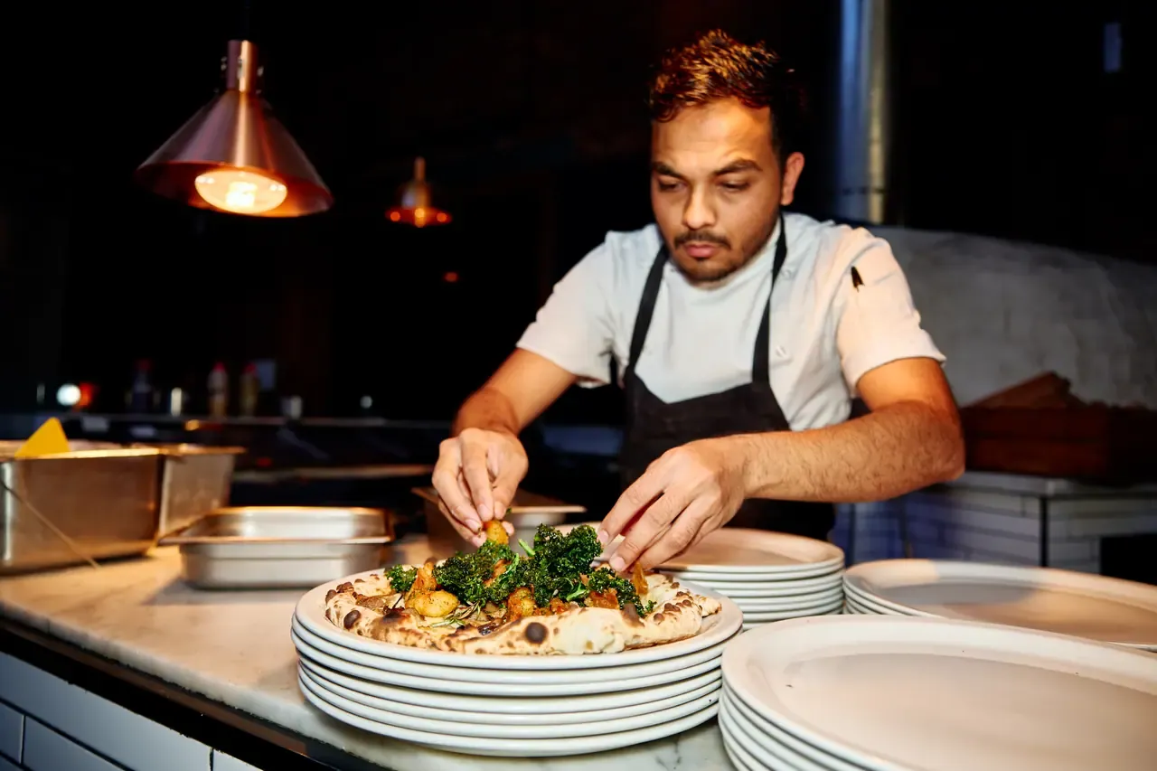 A chef adding greens to a wood-fired pizza at the pass beneath copper lamps at The Epicurean in Red Hill