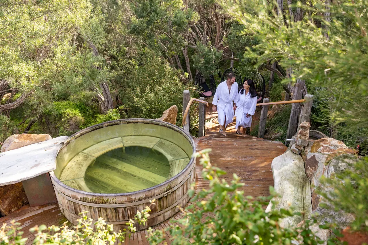 Two bathers in white robes walk a timber boardwalk past a round wooden soaking tub in the bush at Peninsula Hot Springs