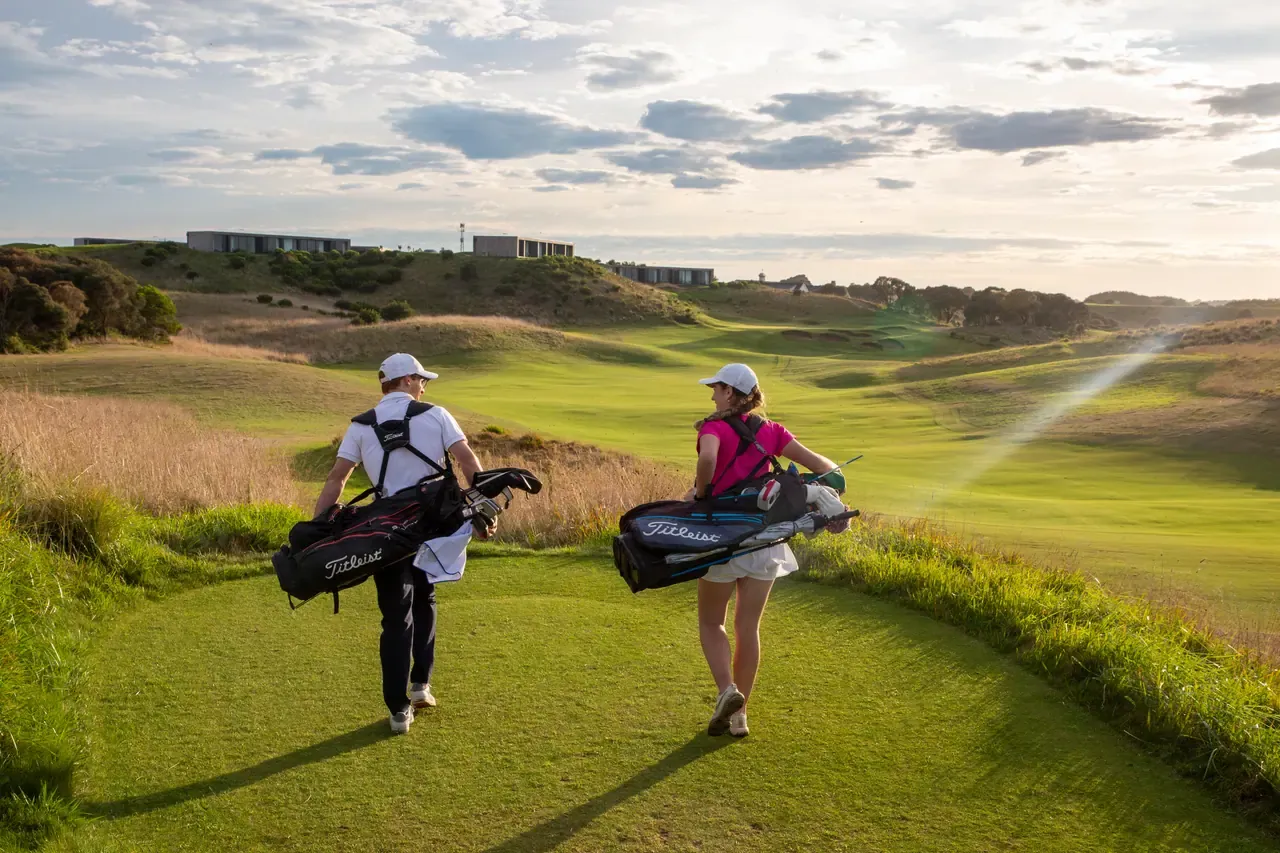Two golfers carrying bags walk toward a rolling fairway at The Dunes Golf Links, with low buildings on the ridge and sun through cloud
