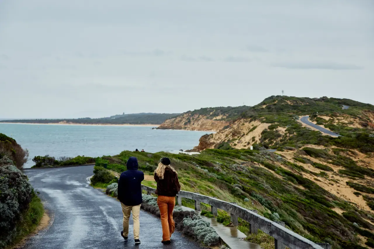 Two walkers in jackets head along a damp coastal road at Point Nepean National Park, with scrubby dunes and a bay ahead.
