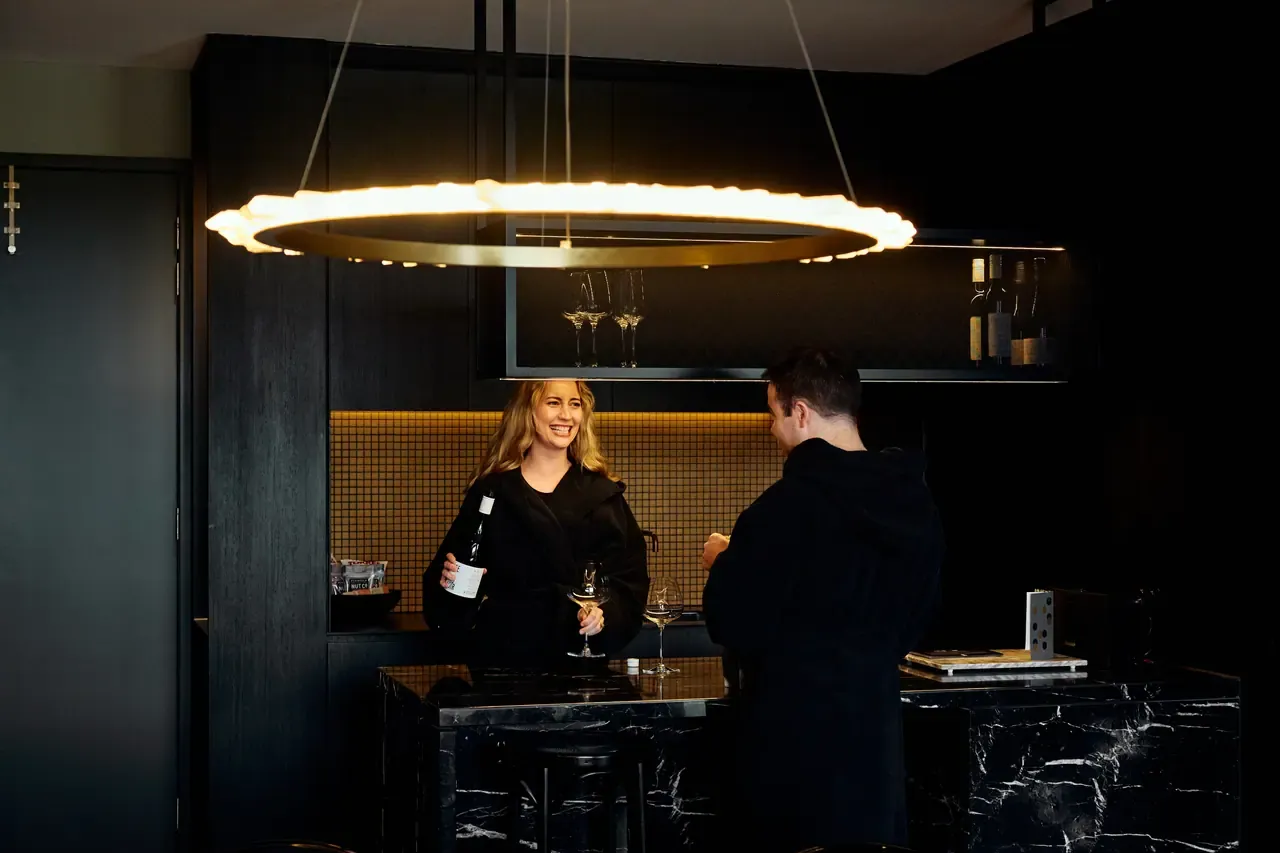 Two guests in dark robes share wine at a black marble bar beneath a glowing ring pendant light in a Jackalope Hotel suite