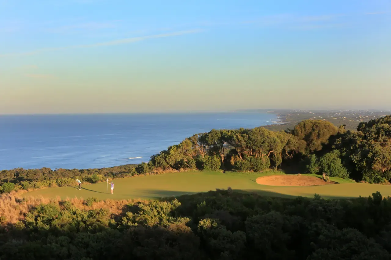 Clifftop green with a bunker at The National Golf Club, two small golfers on it and the coastline curving away beside the sea
