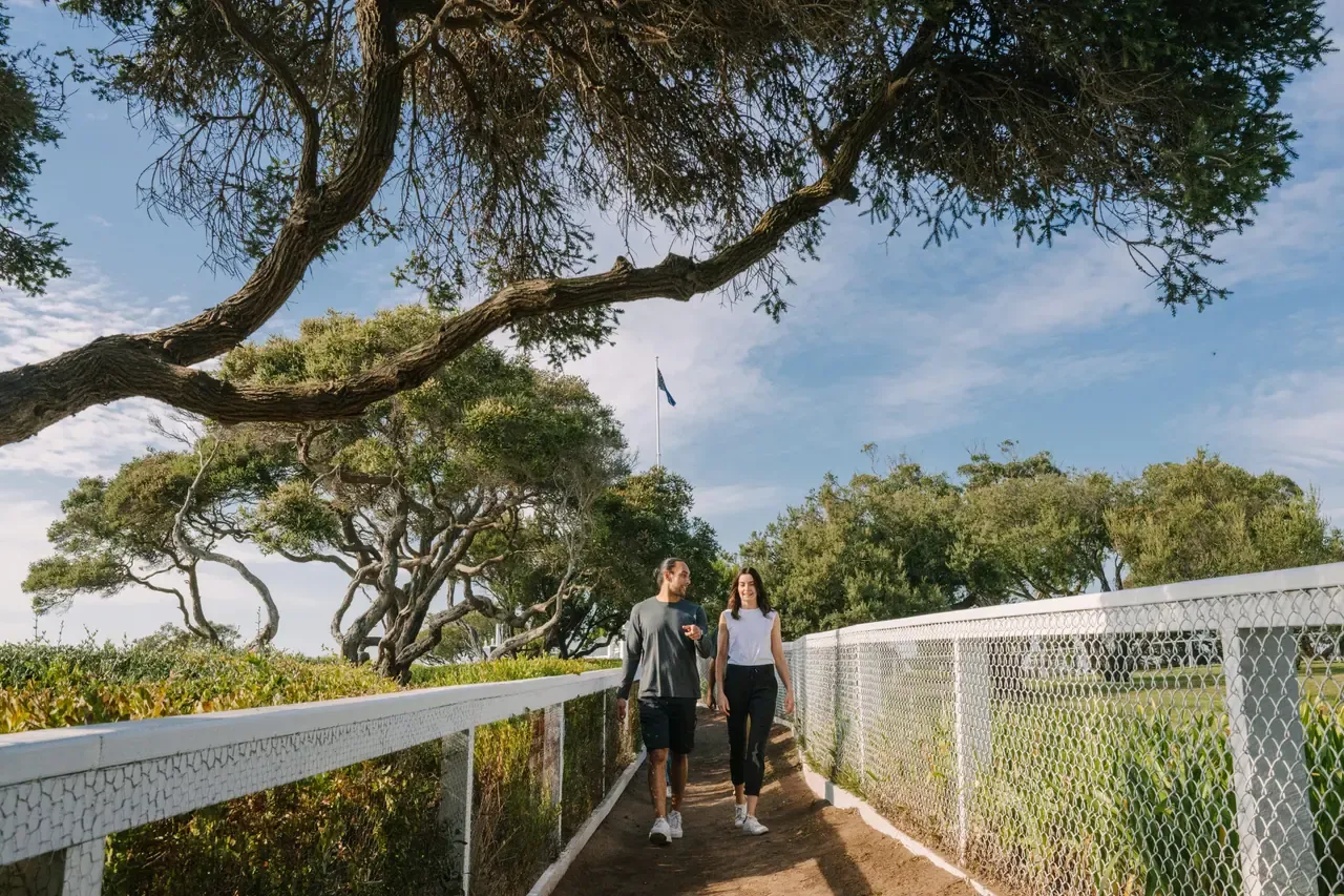 A couple stroll toward the camera on a fenced path under an arching coastal tree on Millionaire's Walk