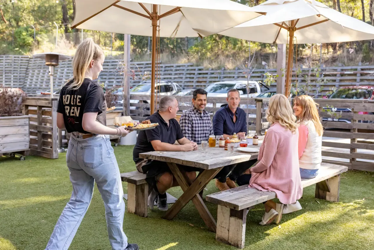 A server brings plates to a group at a picnic table on the lawn under white umbrellas at Red Gum BBQ
