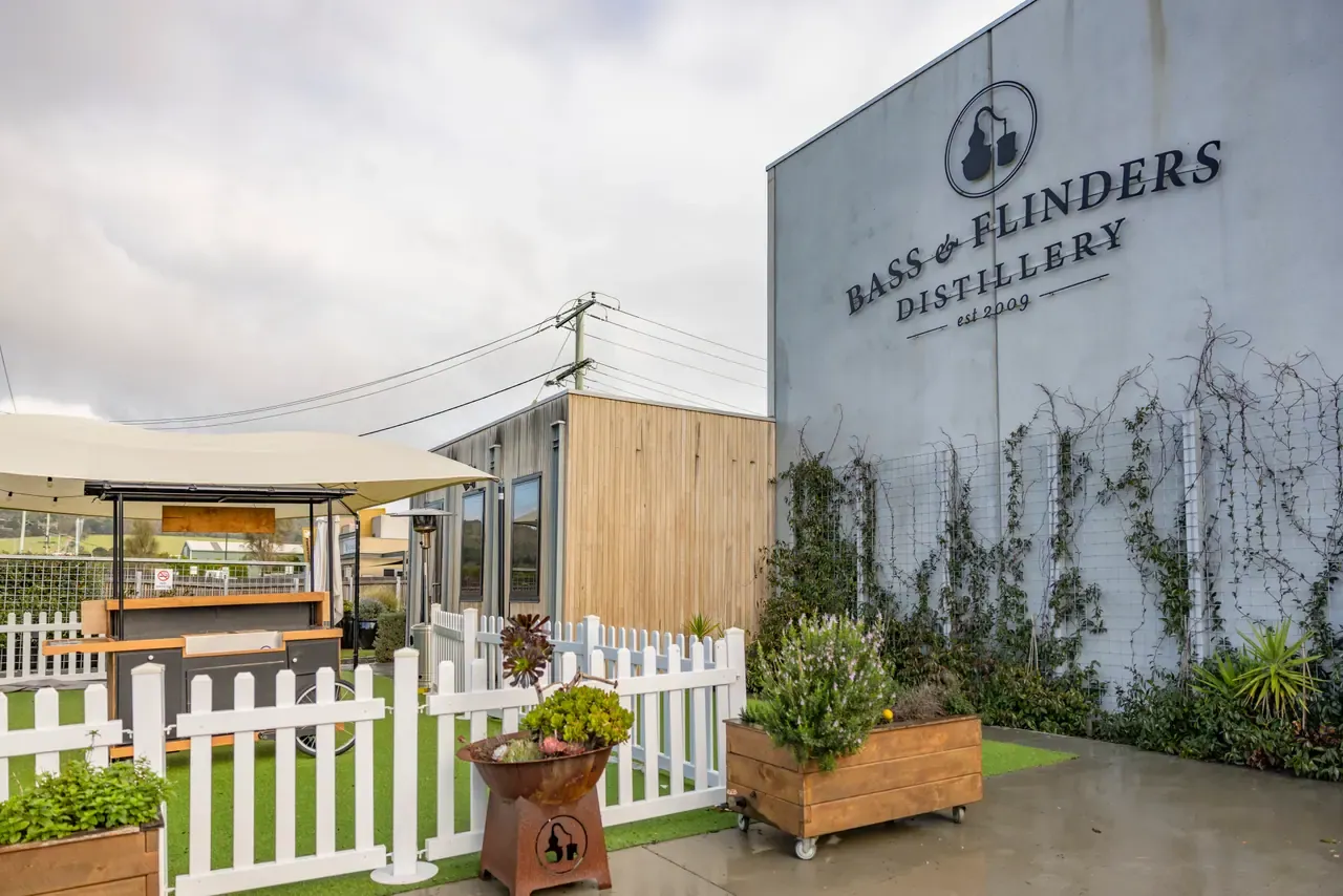 Bass and Flinders Distillery signage on a tall grey wall beside a picket-fenced garden, planter boxes and a small bar