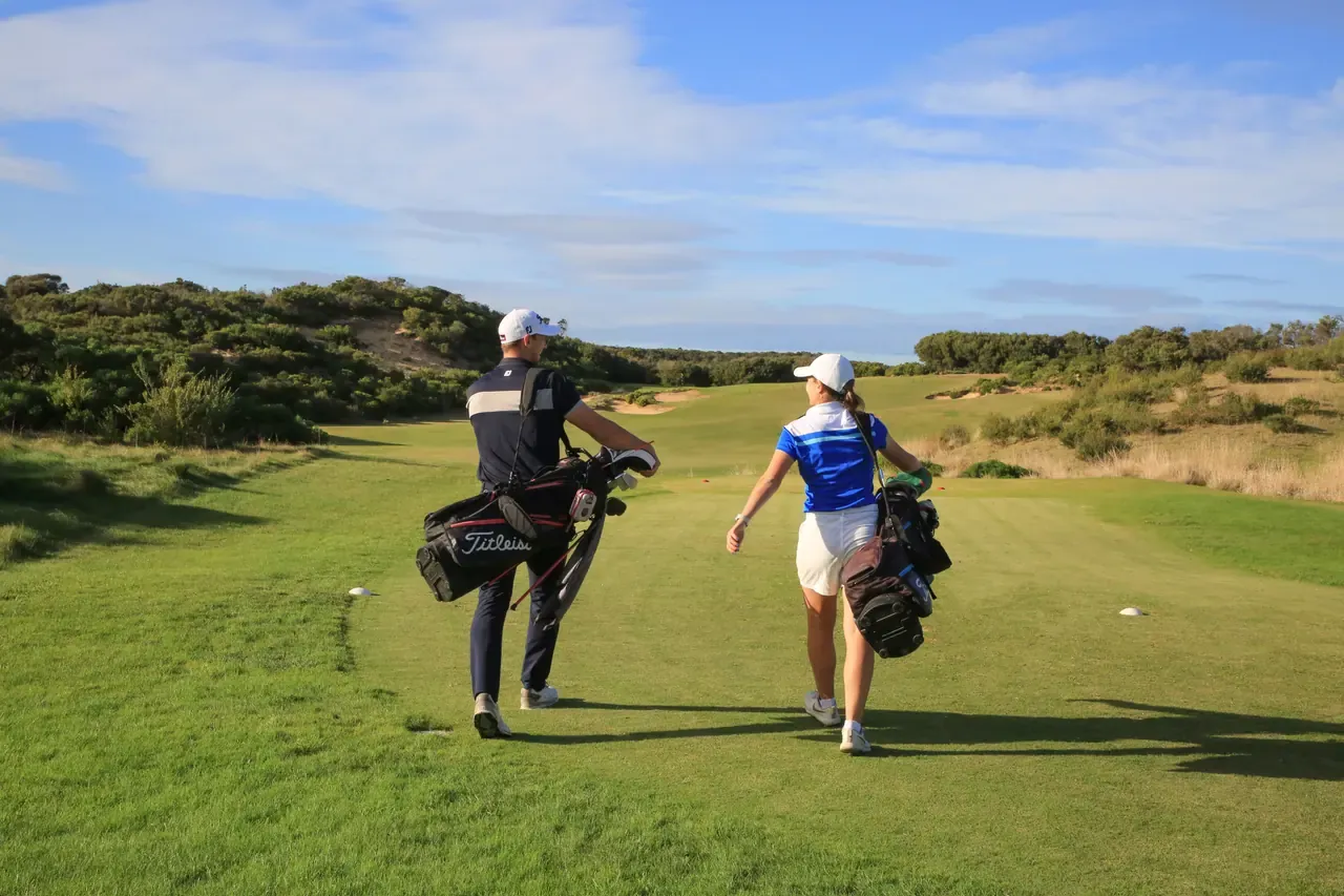 Two golfers carrying bags walk down a wide fairway at St Andrews Beach Golf Course toward sandy dunes and coastal scrub
