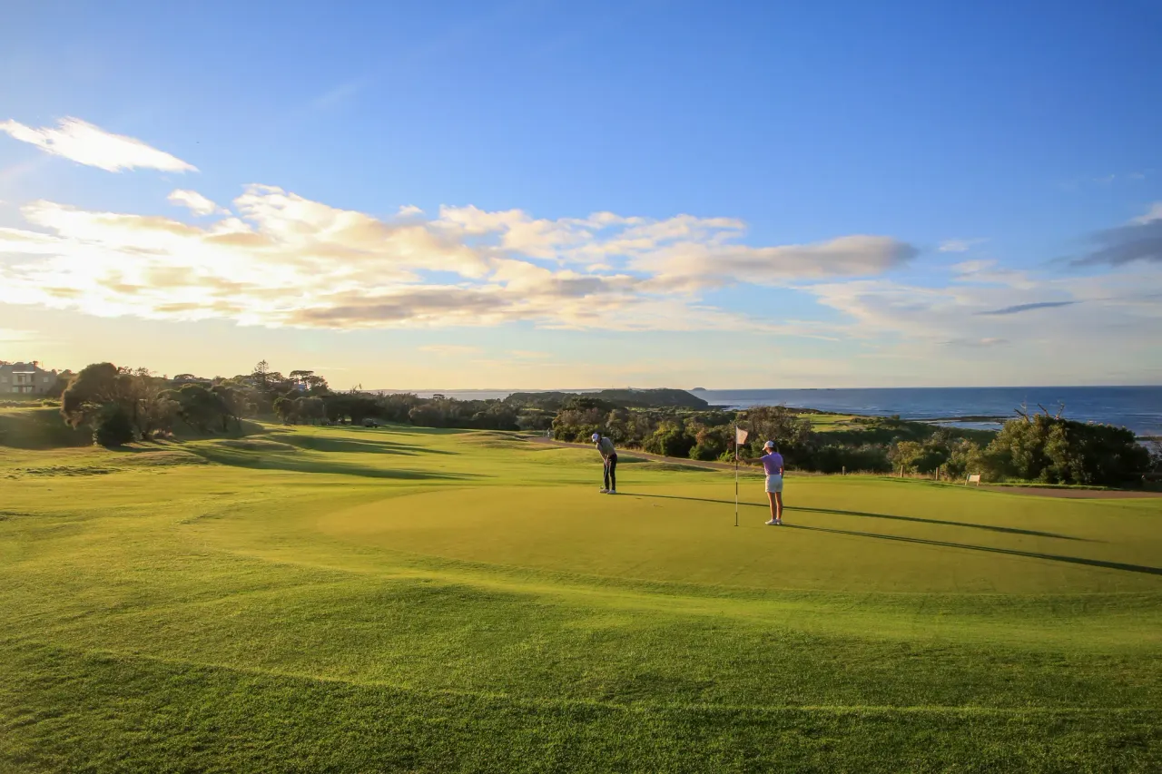 Two golfers on a clifftop green at Flinders Golf Course, with Bass Strait and a low headland beyond under late sun
