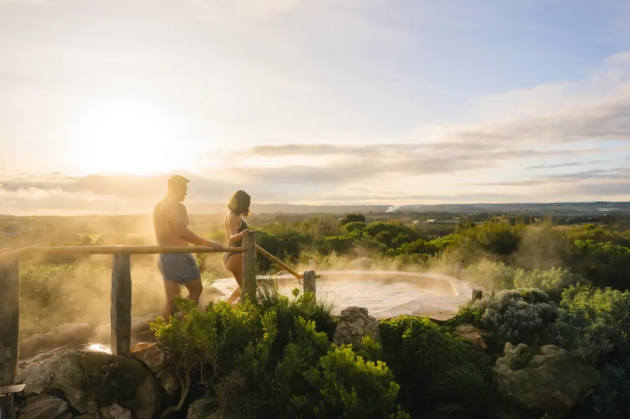Two bathers walk to a steaming hilltop pool at Peninsula Hot Springs as the low sun lights the surrounding bushland