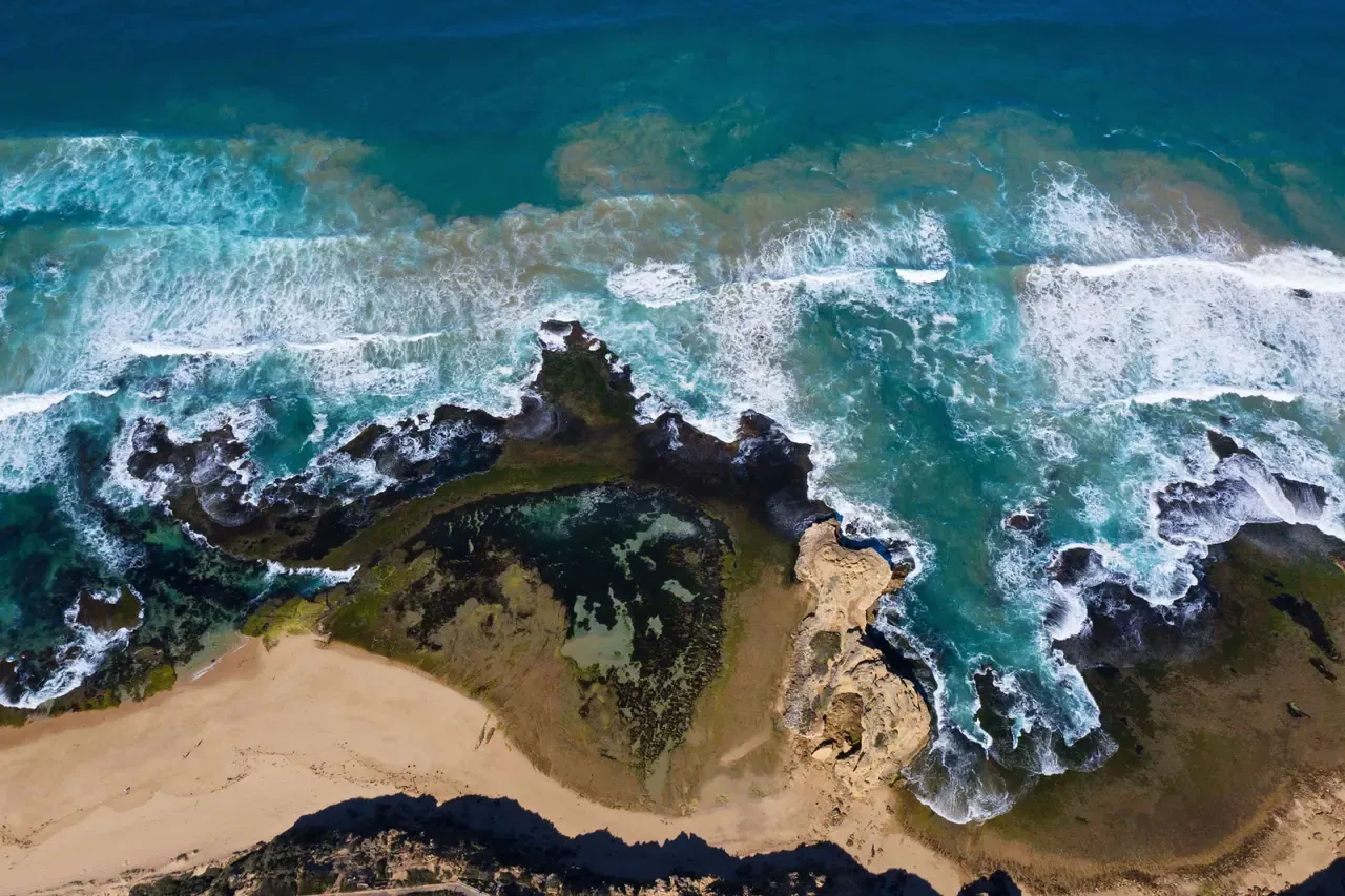 Aerial view of Sorrento Back Beach, where waves wash over a rock platform and rock pool beside a curve of sand below the cliffs
