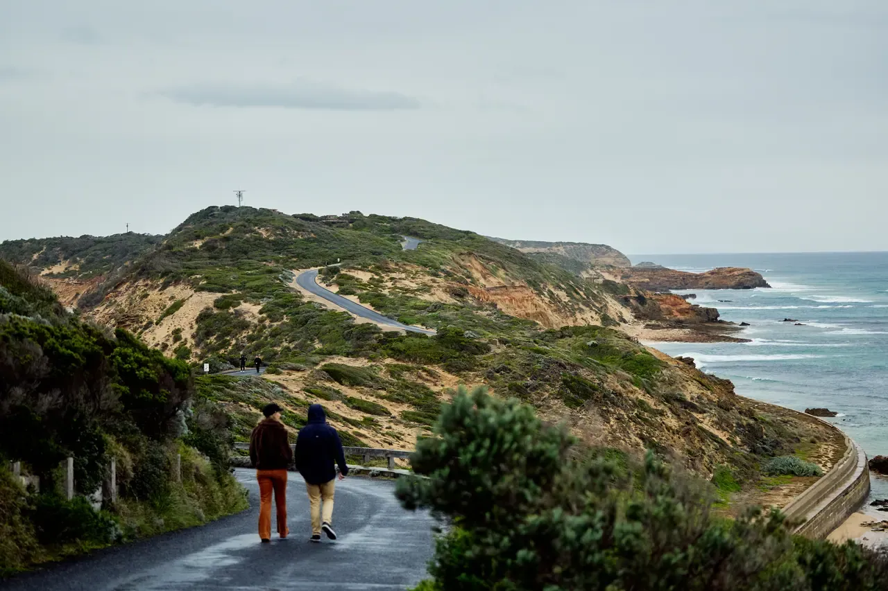 Two walkers head along a sealed road through coastal hills at Point Nepean National Park, with cliffs and rough sea under an overcast sky.