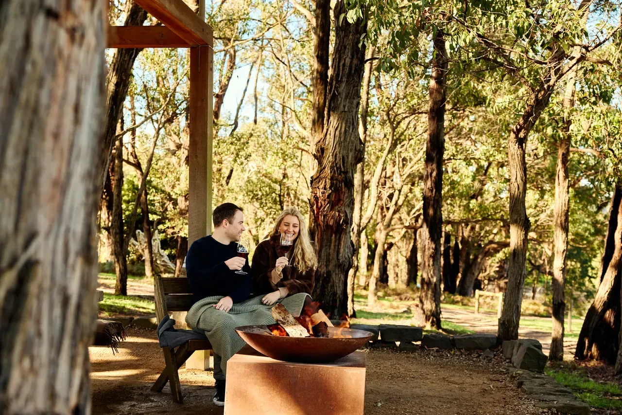 Couple with glasses of red wine on a bench beside an outdoor fire pit among gum trees, a blanket over their knees