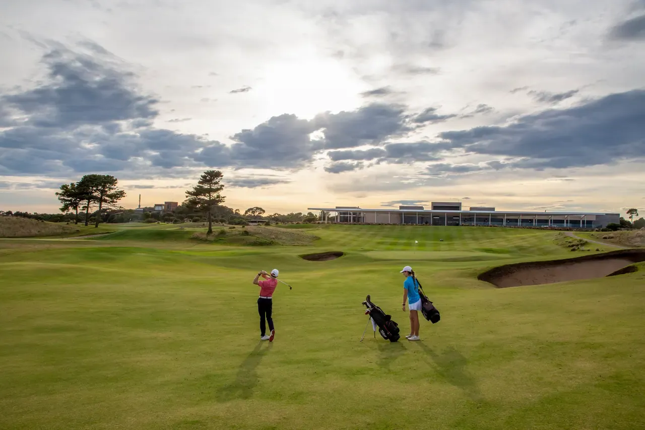 A golfer swings on a fairway at Moonah Links as a partner waits by the bags, the long low clubhouse on the rise beyond
