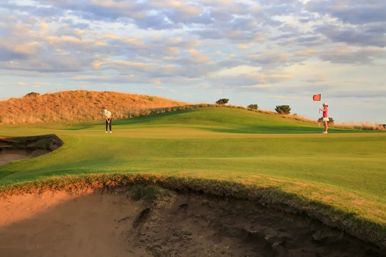 Golfers putting on a green at The Dunes Golf Links, a red flag against grassy dunes and a deep bunker in the foreground