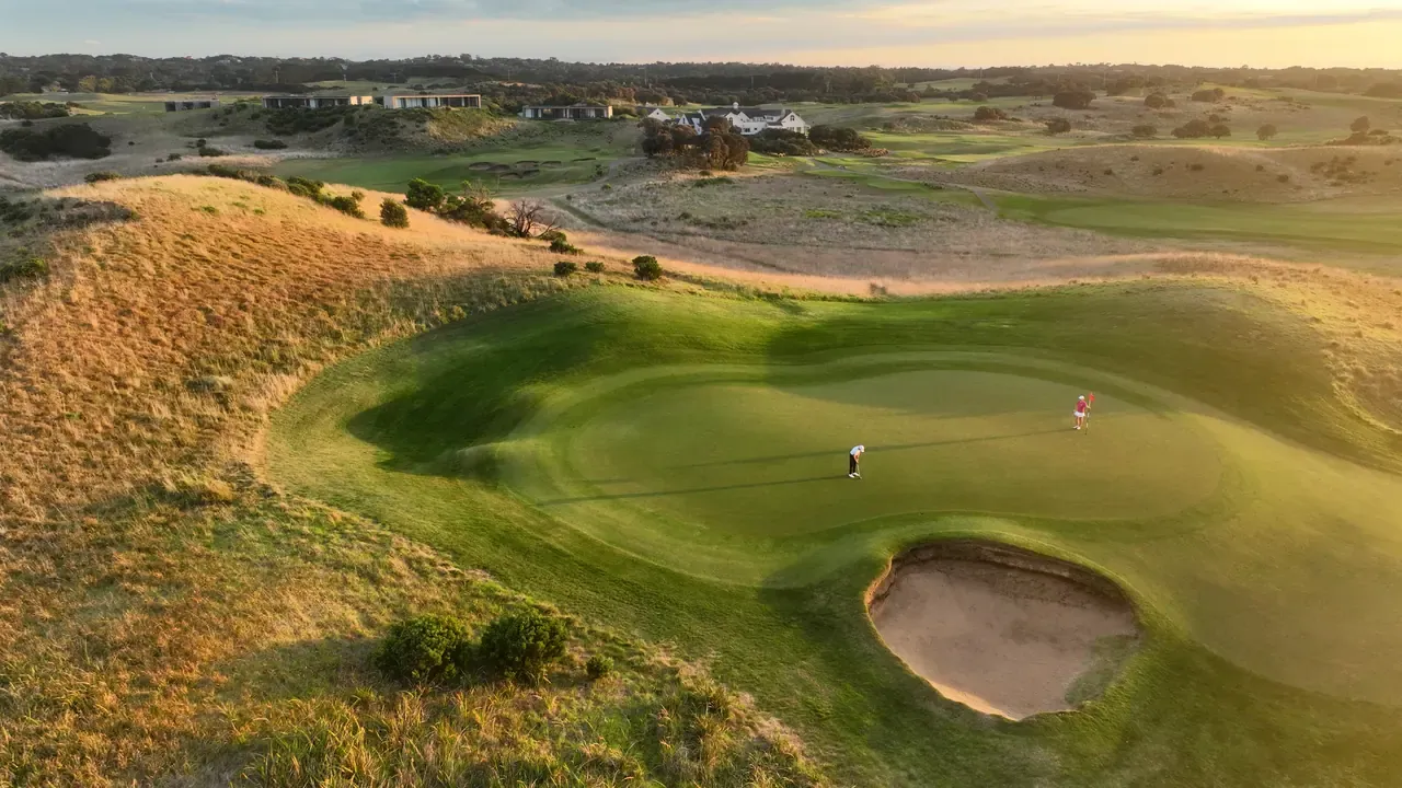 Aerial view of two golfers on a contoured green beside a sand bunker at The Dunes Golf Links in Rye, with golden dune grass in low sun.