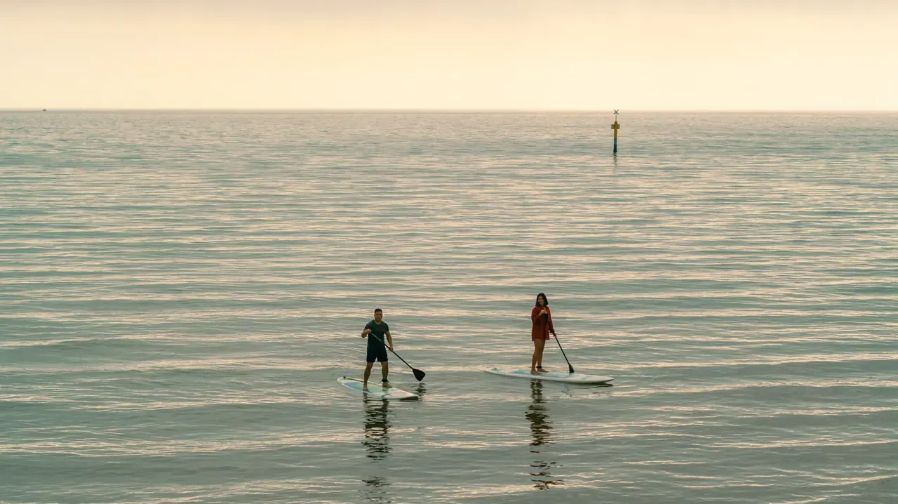 Two stand-up paddleboarders on calm, softly lit water off Mills Beach, with a channel marker in the distance