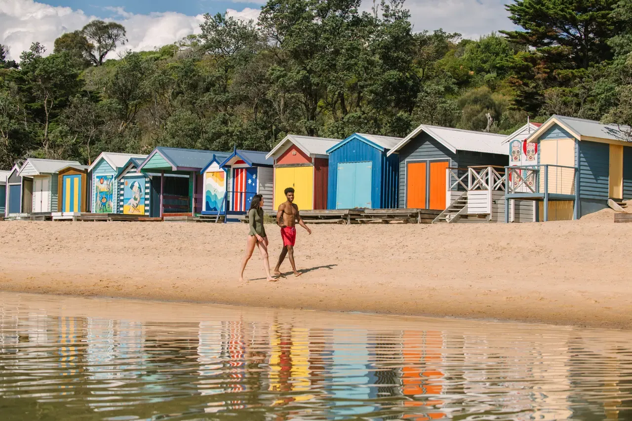 Two people walk along the sand past a row of brightly painted bathing boxes, reflected in the shallows at Mornington
