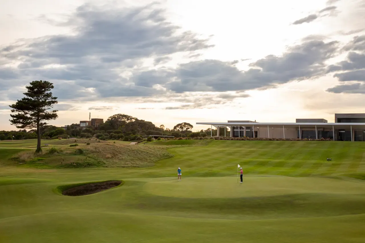 Two golfers on a green beside a pot bunker at Moonah Links, with a lone pine and the long clubhouse under evening cloud