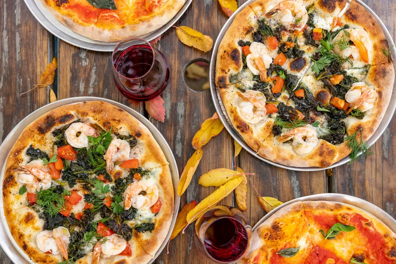 Overhead view of prawn and greens pizzas and a margherita on a timber table, with glasses of red wine and autumn leaves