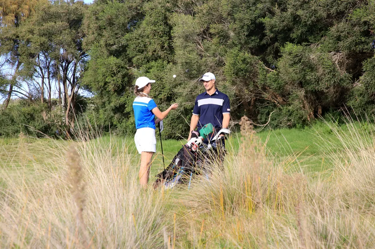 Two golfers chat beside a golf bag in the long grass rough at St Andrews Beach Golf Course, backed by dense coastal trees