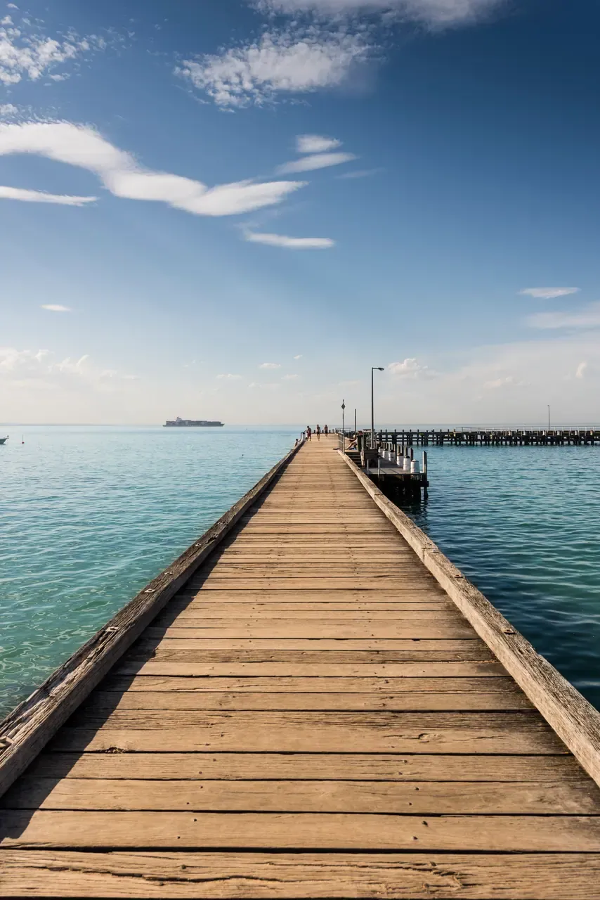 The long timber boardwalk of Portsea Pier leading out over turquoise water, with a cargo ship on the horizon