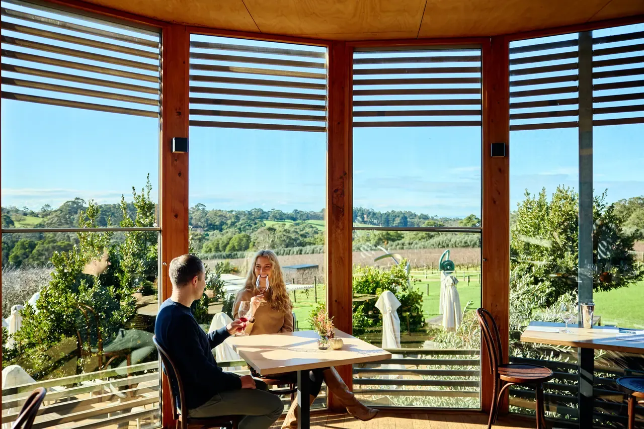 Couple sharing wine at a table inside a timber-framed glass pavilion at Montalto, looking out over vines and bushland